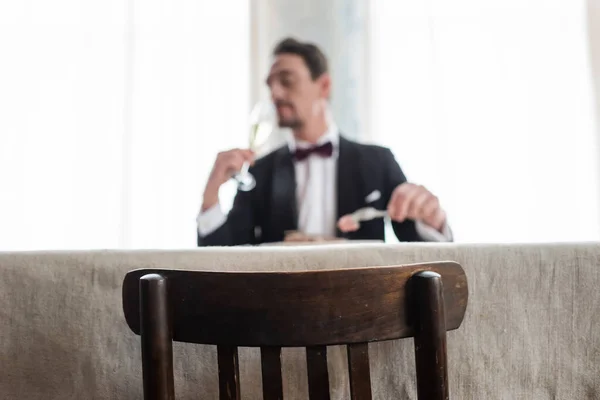 focus on wooden chair, wealthy gentleman in tuxedo enjoying dinner in dining room, natural light