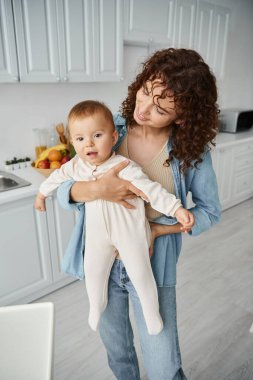 cheerful curly woman holding cute toddler kid in romper in contemporary kitchen at home, happiness clipart