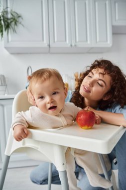Sevgi dolu bir anne, bebek kızına bakıyor. Olgun taze elmanın yanında oturuyor. Mutlu bir ebeveyn.