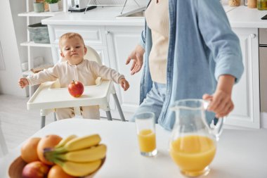 cute kid in baby char looking at ripe apple near mother with jug of orange juice, morning in kitchen clipart
