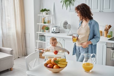 Bir bardak portakal suyu içen kıvırcık kadın mutfaktaki bebek sandalyesinde olgunlaşmış meyvelerle çocuğa bakıyor.