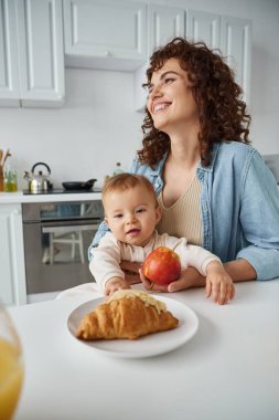 Olgun elmalı, neşeli bir kadın bebek kızına ve lezzetli kruvasana bakıyor. Mutlu kahvaltı.