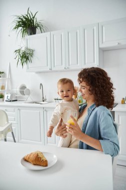 Modern mutfakta kahvaltıda elinde mutlu bir bebek ve bir bardak taze portakal suyu tutan gülümseyen bir kadın.