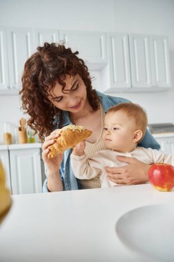 Kıvırcık kadın taze elmanın yanındaki küçük kızına lezzetli kruvasan veriyor, mutfakta kahvaltı.