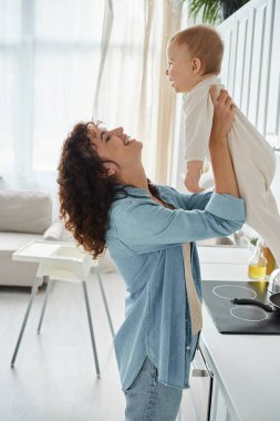 Yeni yürümeye başlayan kızını yetiştiren neşeli bir kadının yan görünüşü modern mutfakta, kaliteli bir zaman.
