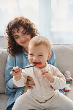 excited little girl playing with teething toy near smiling mother on couch at home, fun and leisure clipart