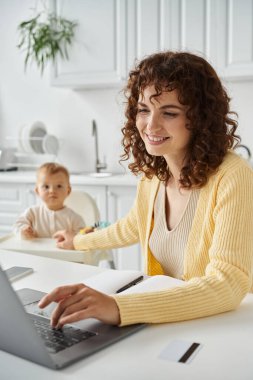 Mutfak, iş ve aile dengesi gibi küçük bir çocuğun yanında dizüstü bilgisayarda çalışan mutlu bir kadın serbest yazar.