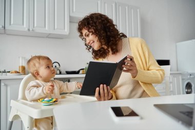 Küçük kızına defterini gösteren neşeli bir kadın mutfaktaki bebek koltuğunda oturan, çalışan bir ebeveyn