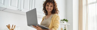 happy woman with wavy hair standing with laptop in modern kitchen, freelancer and housewife, banner clipart