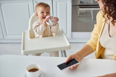 Neşeli çocuk annesinin yanındaki biberondan içiyor. Yanında kahve fincanının yanında boş ekranlı akıllı telefon var.