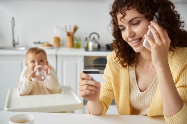 woman with credit card talking on smartphone during online shopping near daughter with baby bottle clipart