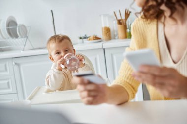 Akıllı telefon ve kredi kartıyla annenin yanındaki biberondan içen küçük kız ön planda bulanık duruyor.