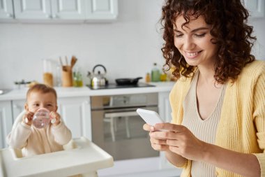 Gülümseyen kadın cep telefonuyla mesajlaşırken küçük çocuk mutfakta bebek şişesinden içiyor.
