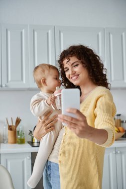Küçük kızıyla cep telefonundan selfie çeken neşeli bir kadın mutfakta bebek şişesi tutuyor.