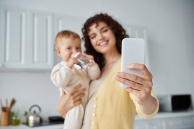 Mutlu anne akıllı telefondan bebek kızıyla selfie çekip mutfakta bebek şişesinden içiyor.