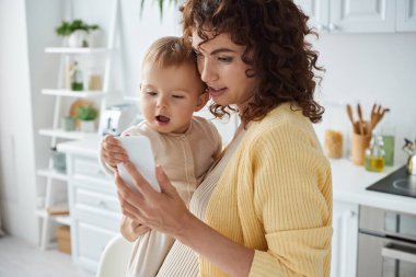Heyecanlı bir çocuk tutan ve interneti karıştıran bir kadın mutfaktaki akıllı telefondan, modern ebeveynlik