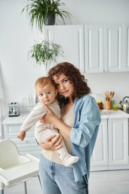 Anne, modern mutfakta sevimli küçük kızını tutarken kameraya bakıyor. Sevgi ve ilgi.