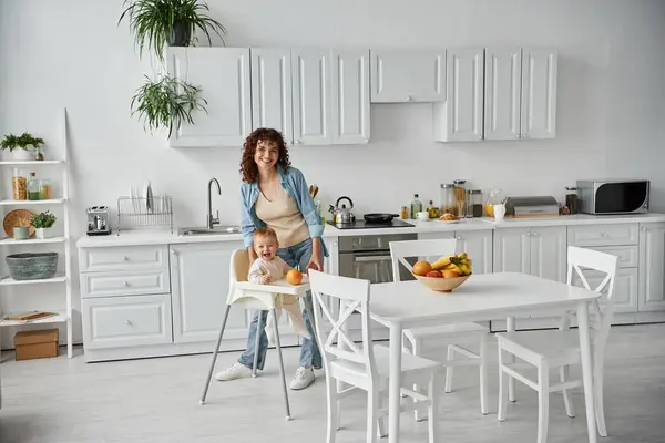 Mutlu anne ve çocuk bebek sandalyesinde beyaz geniş mutfakta olgun meyvelerin yanında kameraya gülümsüyor.
