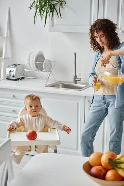 モダンなキッチンで赤ちゃんの椅子と新鮮なフルーツの子供の隣に自然なオレンジジュースを注ぐ母 — ストック写真