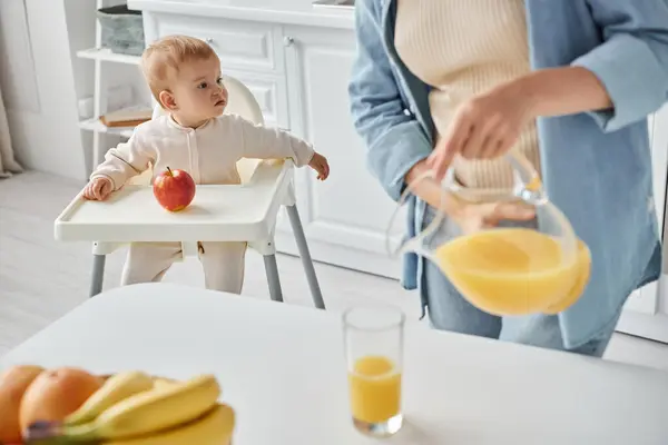 stock image woman pouring fresh orange juice while toddler daughter sitting in baby chair near ripe apple