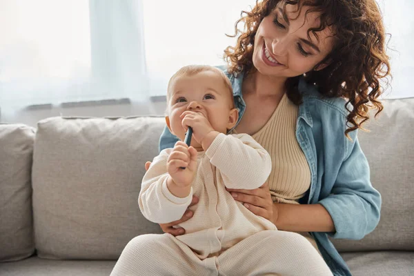 adorable baby chewing teething toy near cheerful mother on couch in living room, leisure and fun