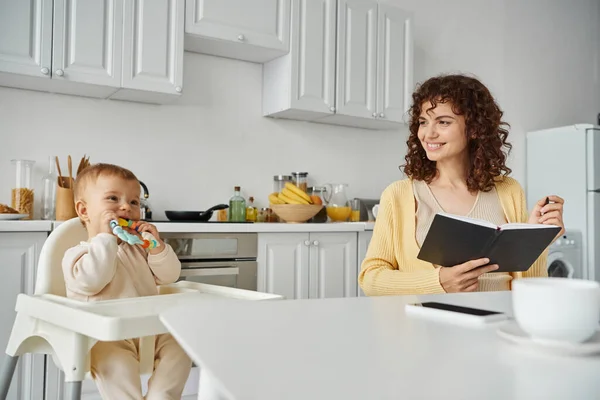 joyful woman with notebook looking at toddler baby with rattle toy while working in kitchen at home