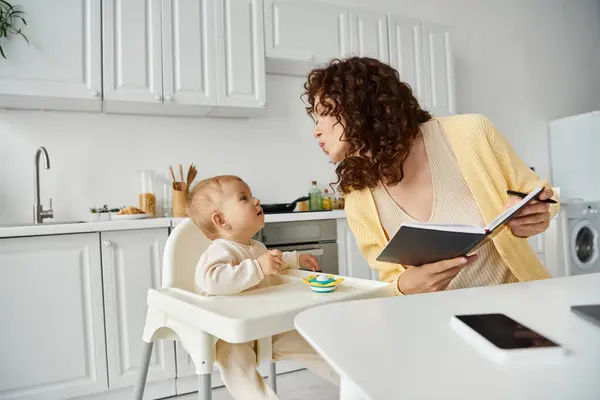 woman with notebook sending air kiss to little daughter sitting in baby chair, working in kitchen