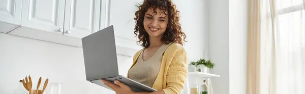 快乐的女人 留着波浪般的头发 站在现代厨房的笔记本电脑旁 自由职业者和家庭主妇 — 图库照片