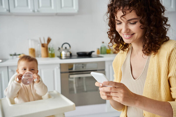 smiling woman messaging on mobile phone while little kid drinking from baby bottle in kitchen