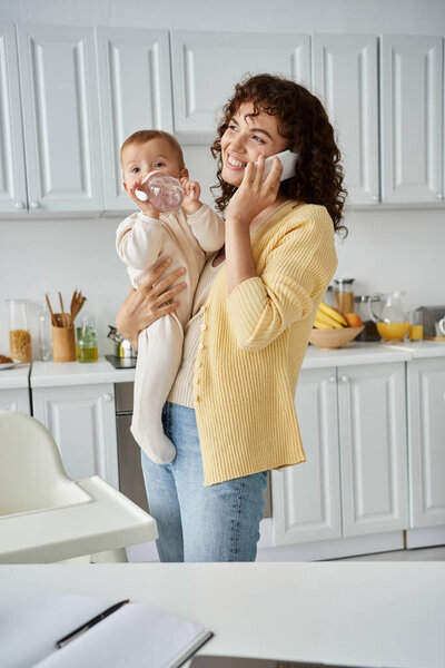 cheerful mother talking on smartphone and holding child drinking from baby bottle in kitchen