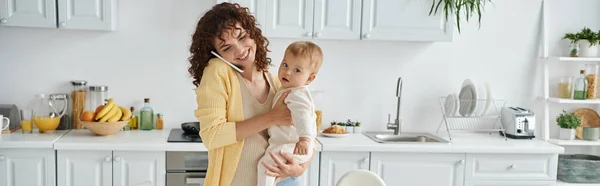 stock image motherhood routine, smiling woman holding little kid while talking on smartphone in kitchen, banner