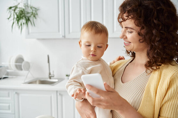 cheerful woman showing mobile phone to toddler girl while standing in modern kitchen, digital age