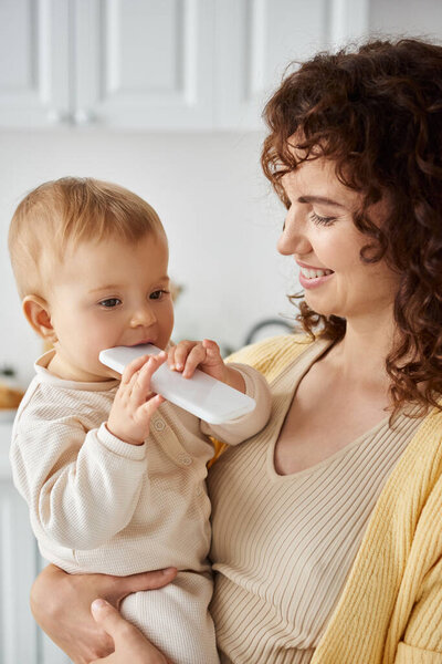 cute baby girl chewing mobile phone near smiling mother at home, blissful childhood in happy family
