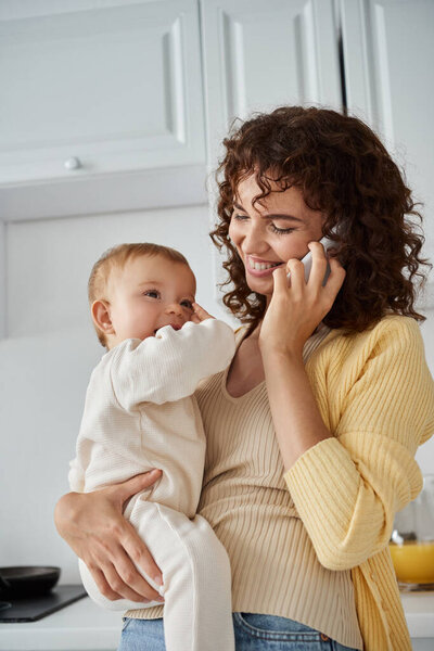 happy woman holding adorable baby girl and talking on smartphone in kitchen, blissful motherhood