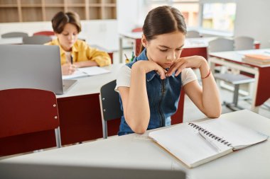 A child with long hair sitting at a desk, focused on writing in a notebook placed in front of her. clipart