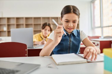 A young girl sits at a desk, holding a pen and notebook, fully engaged in writing or drawing. clipart