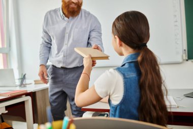 Bir erkek öğretmen, parlak, hayat dolu bir sınıfta küçük bir kızın yanında duruyor. Bir konuşma ya da eğitim seansına katılıyor..