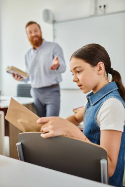 Kız, erkek öğretmeni dinlerken kitap okuyor.