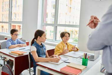 Öğrenmeye dalmış bir grup çocuk, neşeli bir erkek öğretmen parlak bir sınıfa bilgi verirken masalarında dikkatle oturuyorlar..