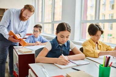 Bir grup çocuk, erkek bir öğretmenin liderliğindeki canlı bir sınıftaki parlak masalarda oturarak öğrenme faaliyetlerine girişiyor..