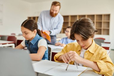 Bir grup çocuk masalarında dikkatle otururken, erkek bir öğretmen onlara parlak ve canlı bir sınıfta talimat veriyor..