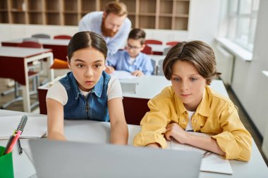 Bir erkek öğretmen, parlak, canlı bir sınıfın masalarında oturan bir grup çocuğa talimat veriyor..