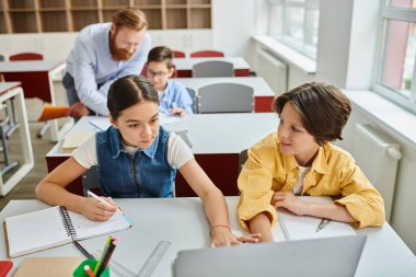 A group of children sit at desks in a bright classroom, listening attentively to a man teacher instructing them. clipart