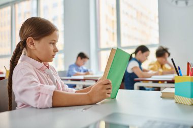 Genç bir kız, parlak bir sınıf ortamında, bir kitaba dalmış bir masada oturuyor..