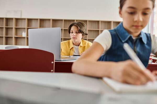 A young boy sits at a desk in a bright classroom, focused on laptop computer