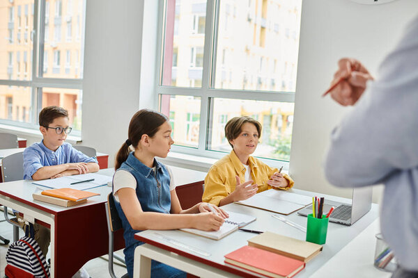 Группа детей, увлеченных учебой, сидит за письменными столами, в то время как живой учитель мужского пола делится знаниями в яркой обстановке класса.