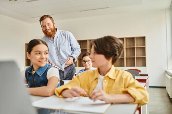 A group of children sit attentively at desks in a brightly lit classroom, engrossed in the computer lesson being taught by their male teacher.