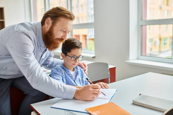 A man sitting at a desk, teaching a young boy in a vibrant classroom setting.