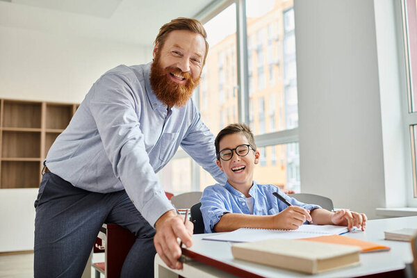 A man standing next to a boy at a desk, engaged in a learning activity in a vibrant classroom setting.