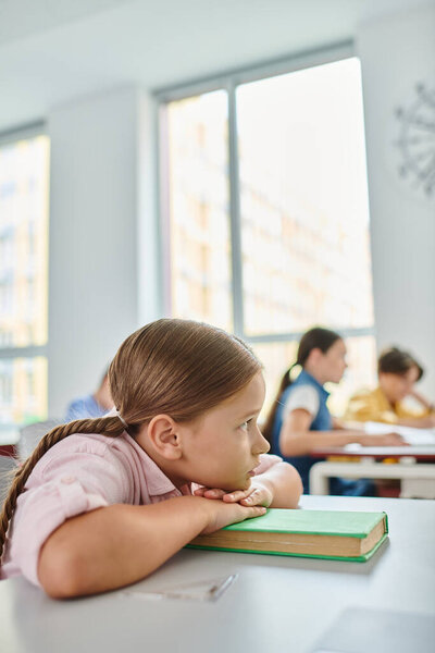 A young girl with pigtails sitting at a desk, completely engrossed in reading a book in lively classroom.